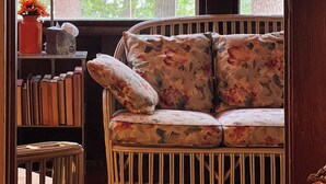 A cozy room with a wicker chair, a floral sofa, a table with a book, a lamp, and a window with a view of trees. - Historic Glen Lake Cottage with Private Waterfront Access (Glen Arbor)