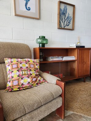 Ein gemütliches Wohnzimmer mit einer Couch, einem gemusterten Zierkissen, einem Holztisch mit einer grünen Lampe, einem Bücherregal und gerahmten Bildern an der Wand.