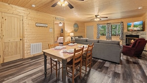 A wooden cabin interior with a dining table, chairs, a sofa, and a television.
