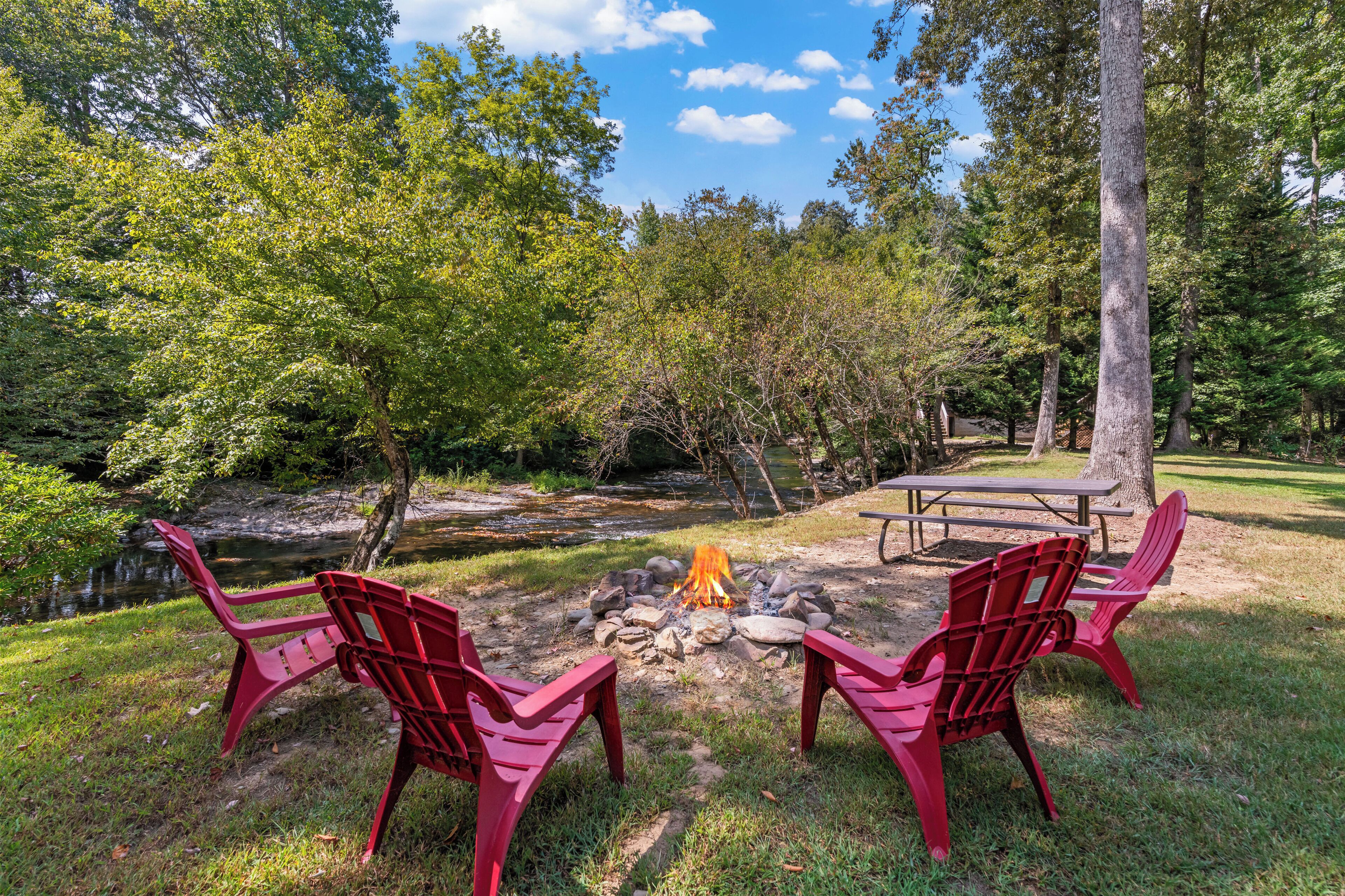 A campsite with a fire pit, two red chairs, a picnic table, and a river in the background.