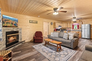 Cozy living room with fireplace, comfortable seating, and a television. Features a kitchen area and wooden ceiling with a ceiling fan.