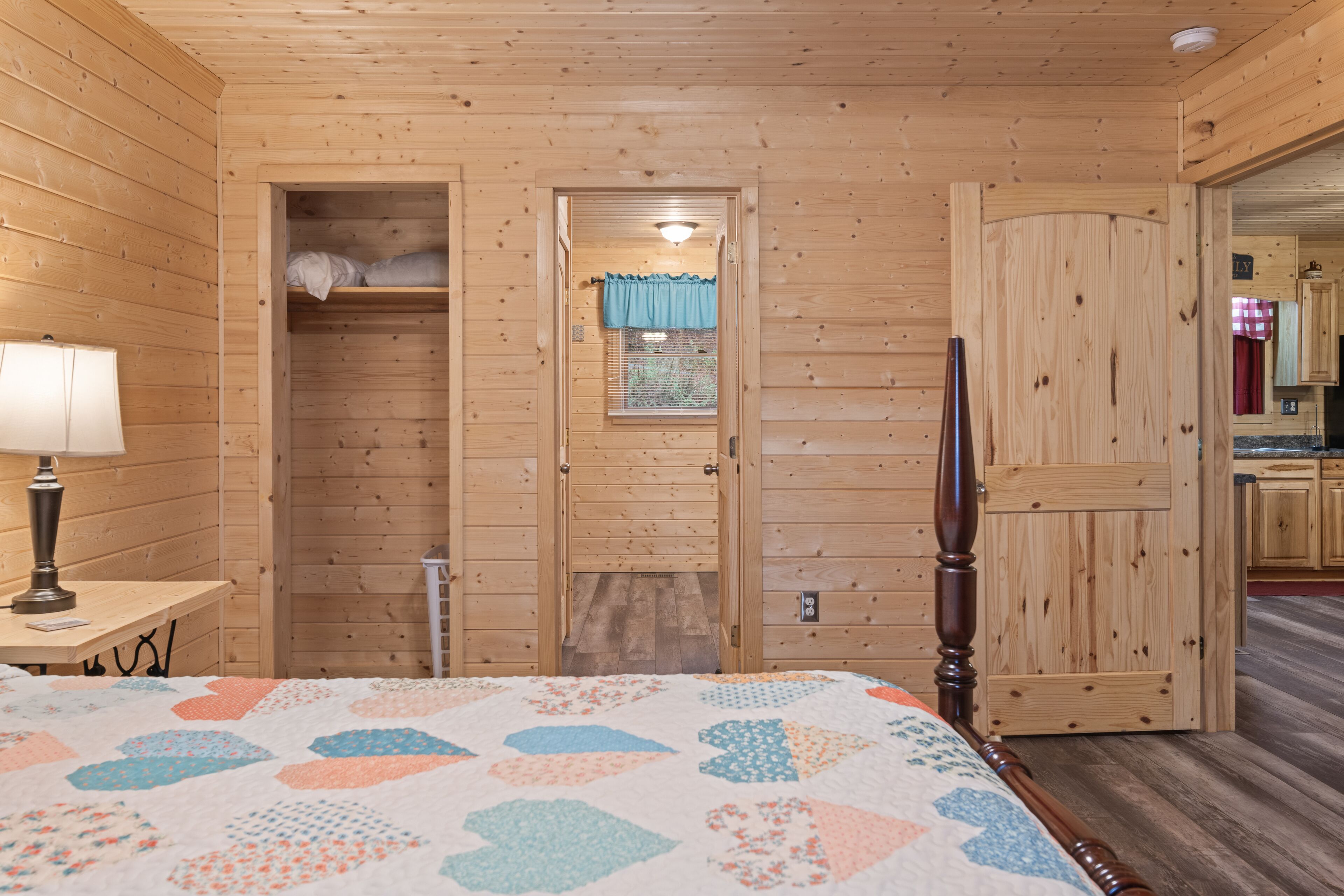 Ein Zimmer einer Holzhütte mit einem Bett, einem Nachttisch, einem Fenster mit Vorhängen und einer Tür zu einer Küchenzeile.