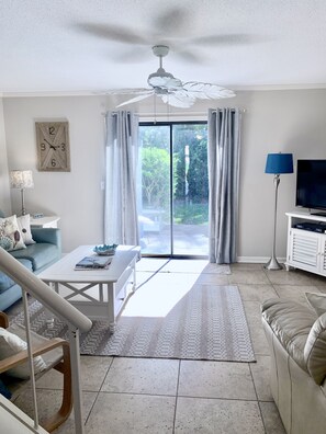 Un salón con sofá blanco, una mesa de centro, televisor, ventilador de techo y vistas a través de un ventanal.