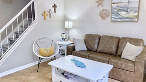 Ein Wohnzimmer mit Treppe, einem weißen Sessel, einem braunen Sofa, einem weißen Couchtisch und Wanddekorationen, darunter ein Seestern und ein Gemälde von Booten.