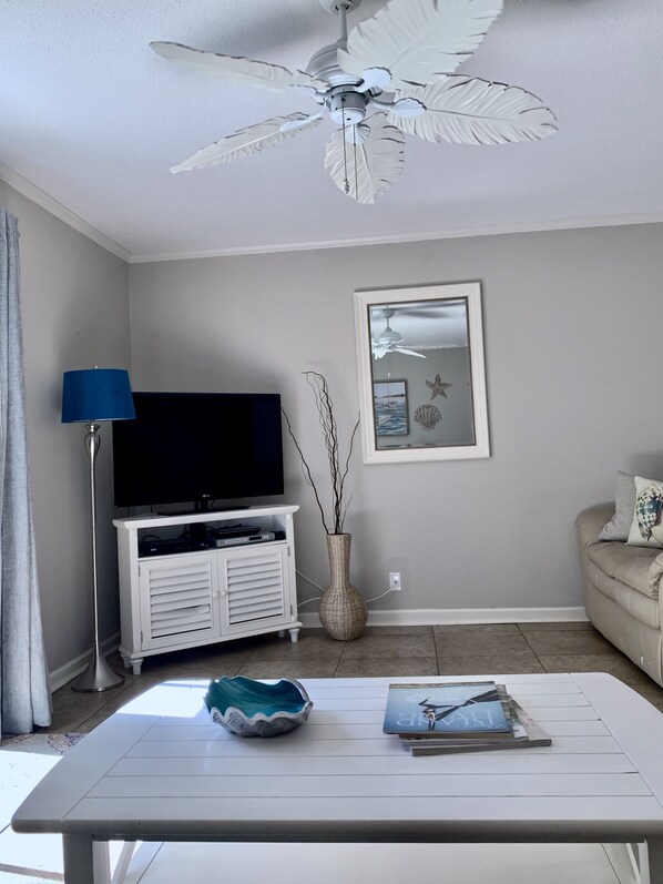 Una sala de estar con ventilador de techo, un televisor de pantalla plana sobre un mueble blanco, un jarrón con ramas, un cuadro enmarcado y un sofá color beige.