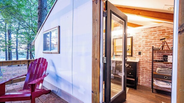 En stuga med en träterrass, en röd Adirondack-stol, en glasdörr som leder till ett badrum med en vägg av tegelsten och ett teleskop på en hylla.