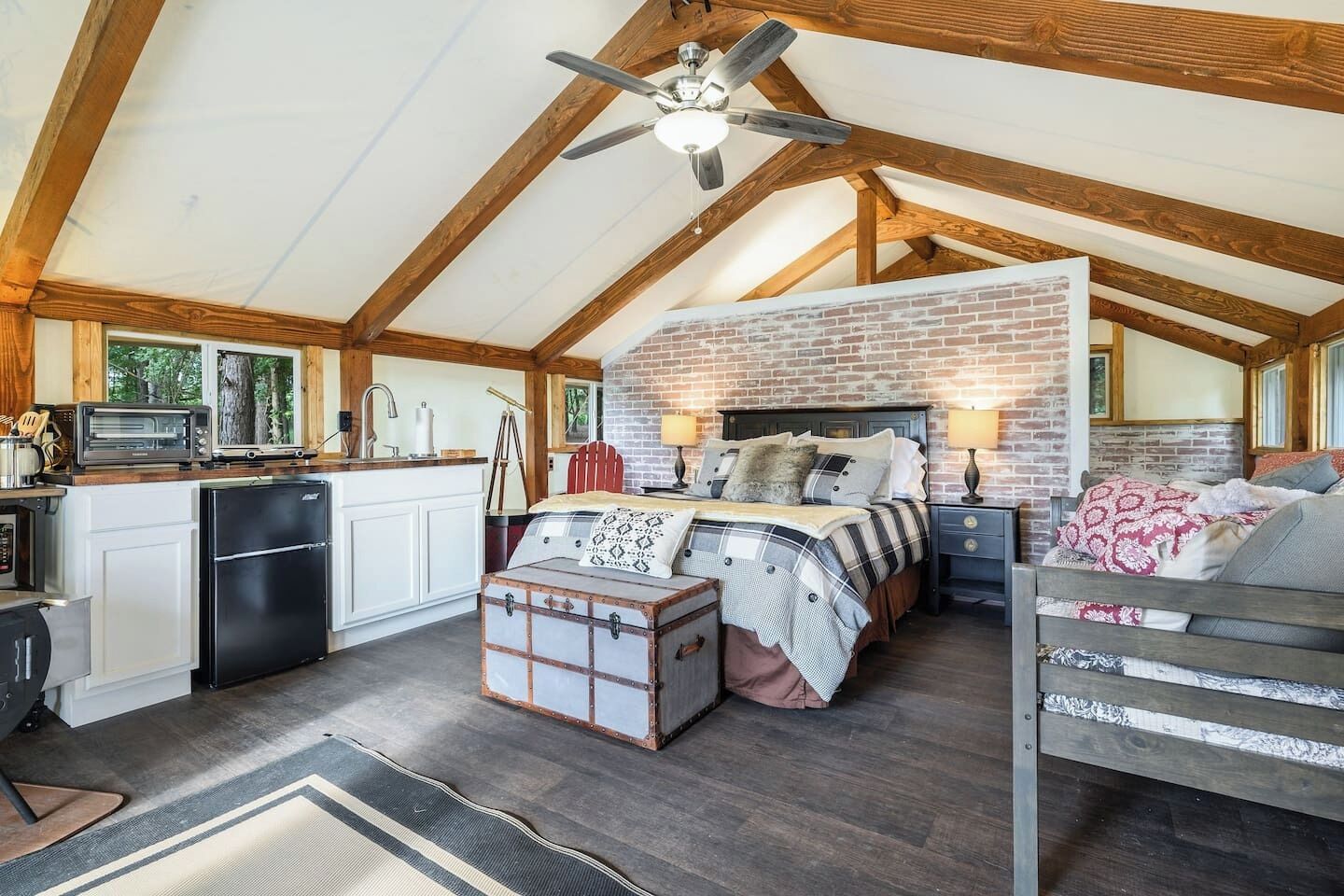 A loft-style bedroom with a brick wall, a bed with multiple pillows, a nightstand, and a dresser.