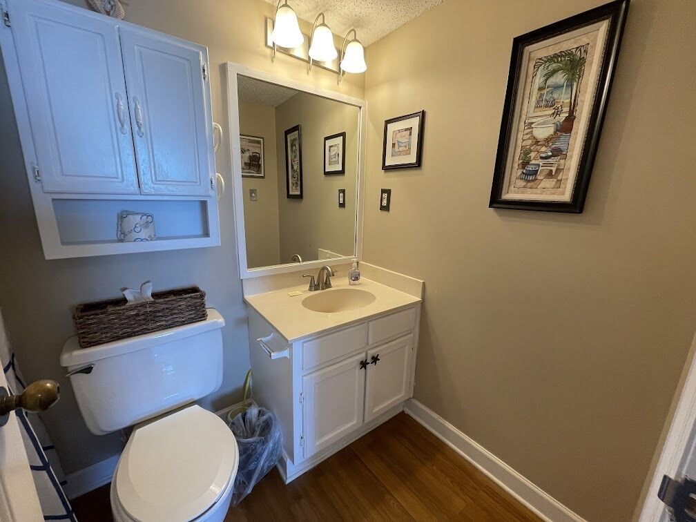 A bathroom with a toilet, sink, and cabinet, featuring a mirror and framed artwork on the wall.