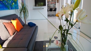 Ein modernes Wohnzimmer mit einem grauen Sofa, bunten Kissen, einem Glastisch und einer Vase mit Blumen.