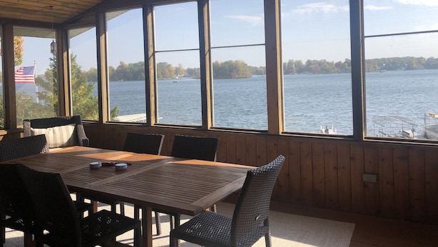 Ein überdachter Essbereich im Freien mit Tisch und Stühlen, große Fenster mit Blick auf einen See und Bäume.