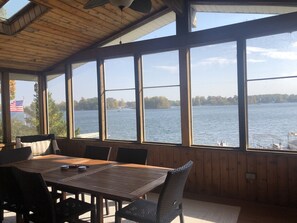 A covered outdoor dining area with a table and chairs, large windows offering a view of a lake and trees. - Magician Lake Life at its best! (Dowagiac)