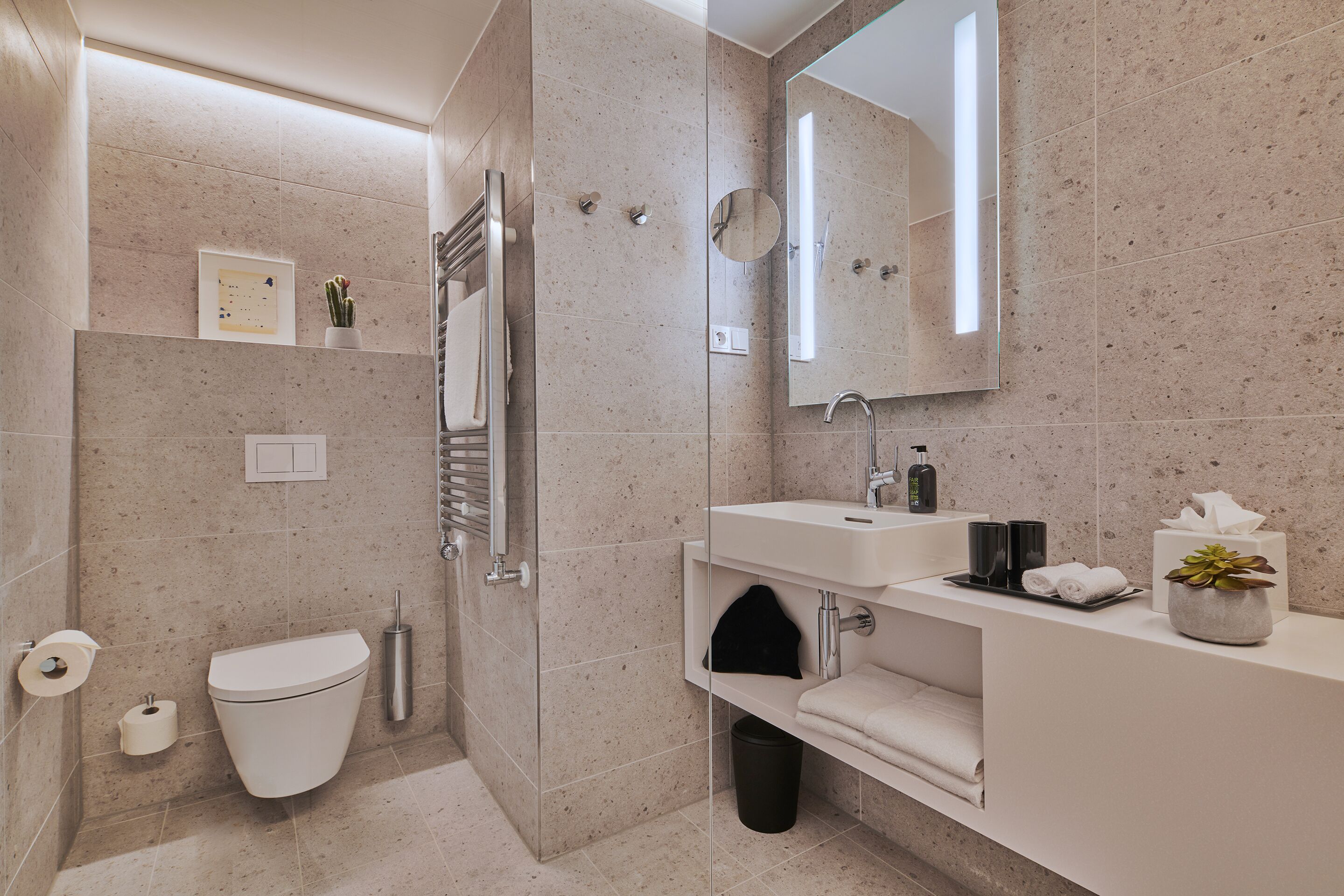 Un baño moderno con inodoro, bidé y zona de ducha. Hay un lavabo con un espejo encima y un estante con toallas y artículos de aseo.