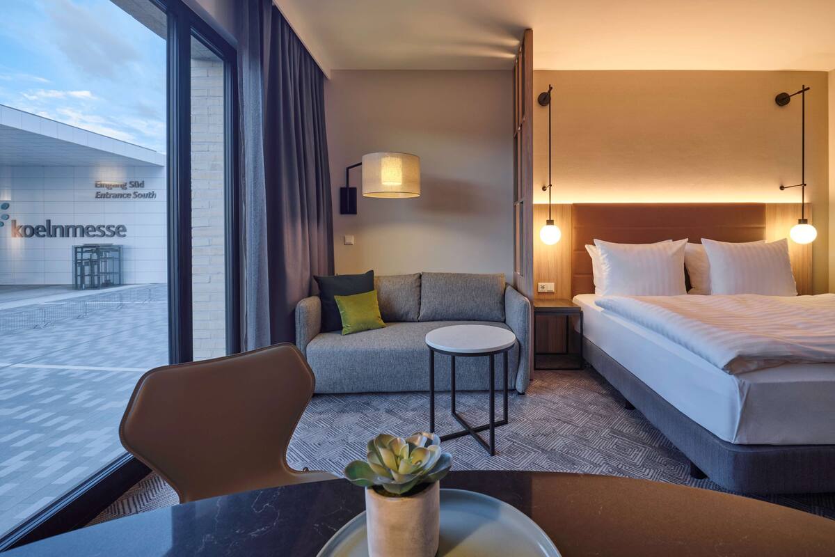 Habitación de hotel con cama, sofá, una mesita con planta y vista al acceso del edificio.