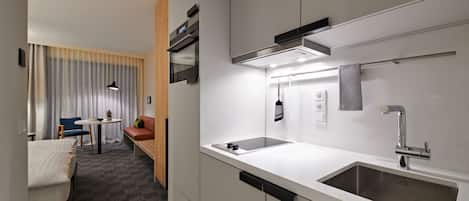 A modern hotel room with a kitchenette, a bed, a seating area with a table and chairs, and a large window with curtains.