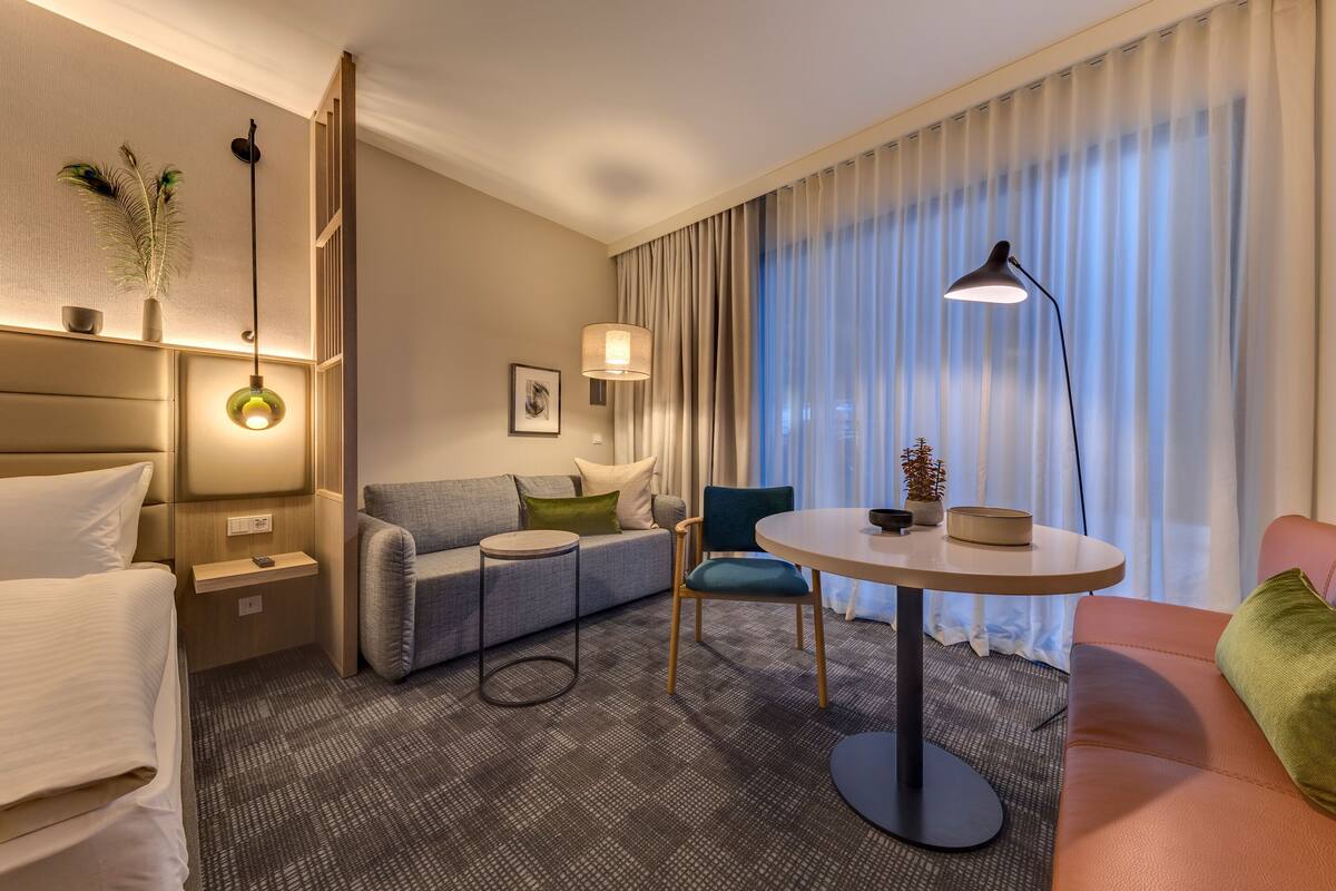 Una habitación de hotel moderna con una cama, un sofá, una mesita y una silla. Hay una lámpara sobre la mesa y una planta en la pared.