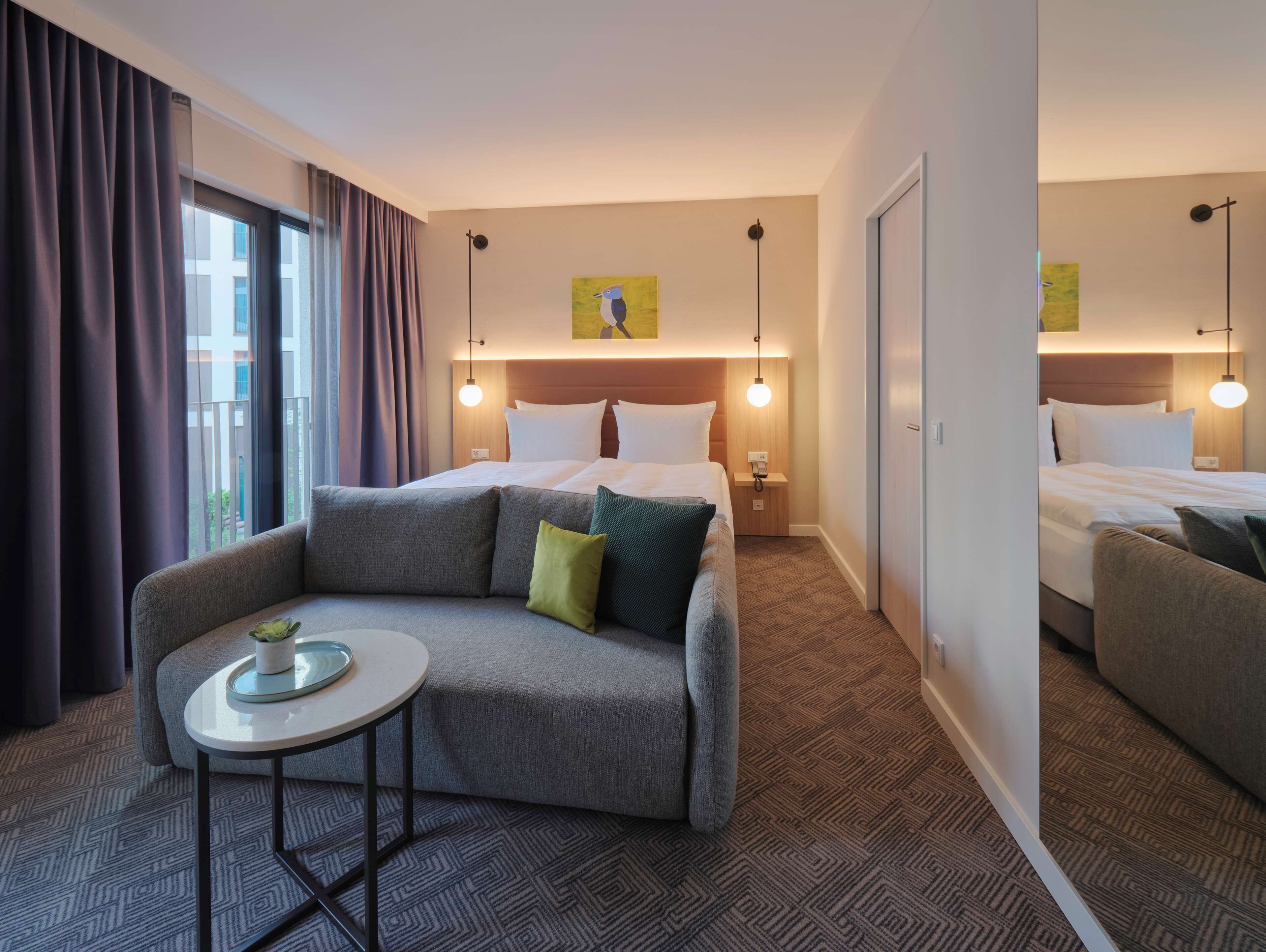 Habitación de hotel con sofá, una mesita, cama con ropa blanca, mesita de noche y un cuadro en la pared.
