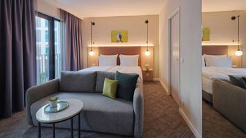 Habitación de hotel con sofá, una mesita, cama con ropa blanca, mesita de noche y un cuadro en la pared.