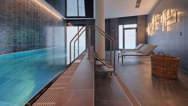 A modern indoor swimming pool with clear glass walls, a metal staircase, lounge chairs, and a woven basket on the floor. - Adina Apartment Hotel Cologne (Cologne)