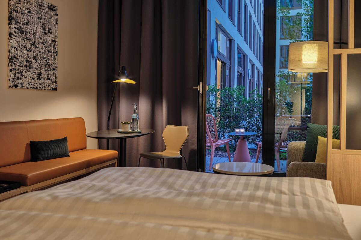 Habitación de hotel con cama, sofá, sillón, una mesita de noche con lámpara y vistas al exterior.