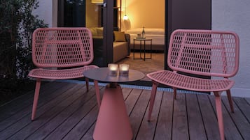 Una terraza de madera con dos sillas de malla rosa y una mesita, una puerta de cristal que da a una habitación con cama y lámpara, y una mesita con una botella y un bol.