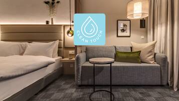 Una habitación de hotel moderna con una cama, un sofá gris, una mesita redonda y una lámpara de pared.