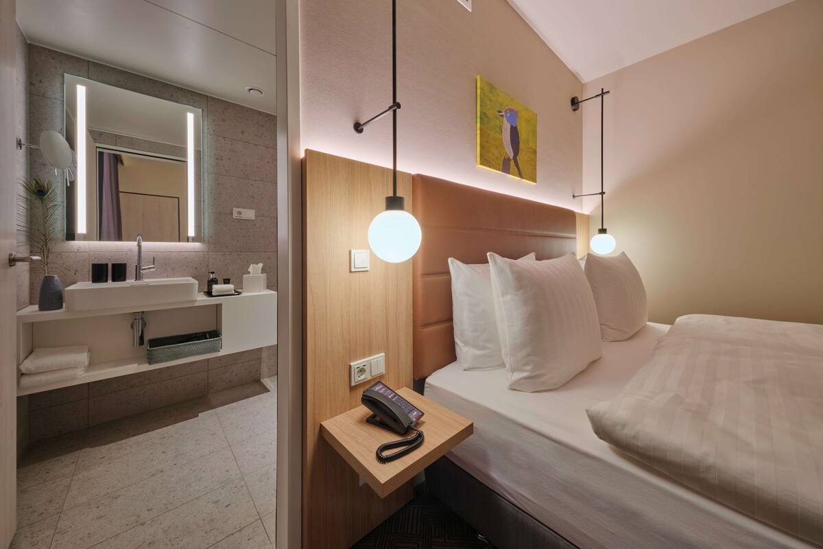 Una habitación de hotel moderna con una cama grande, una mesita de noche con teléfono, un espejo y un estante pequeño con artículos de aseo.