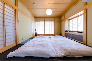 A traditional Japanese room with tatami flooring, a futon bed, sliding doors, and a view through a window with a wooden frame. - iine (Fujikawaguchiko)