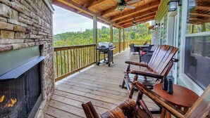 Una terraza de madera con un muro de piedra, chimenea, mobiliario exterior y vistas al bosque.