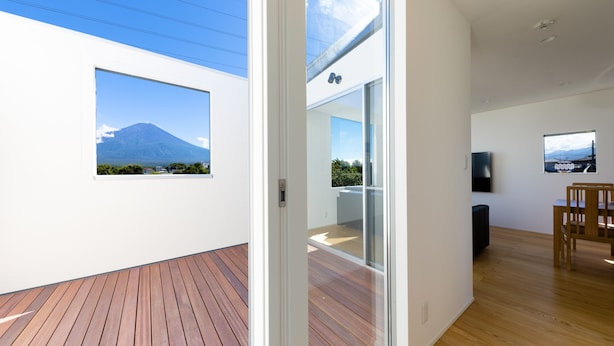 A modern interior with a wooden deck, a glass door leading to a balcony with a mountain view, and a dining area with a TV. - Frame View Mt.Fuji Villa East (Fujikawaguchiko)