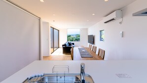 A modern kitchen with a built-in dishwasher, a dining area with a long table and chairs, and a large window offering a view of the outdoors. - Frame View Mt.Fuji Villa East (Fujikawaguchiko)