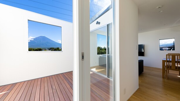 A modern interior with a wooden deck, a glass door leading to a balcony with a mountain view, and a dining area with a TV. - Frame View Mt.Fuji Villa West (Fujikawaguchiko)