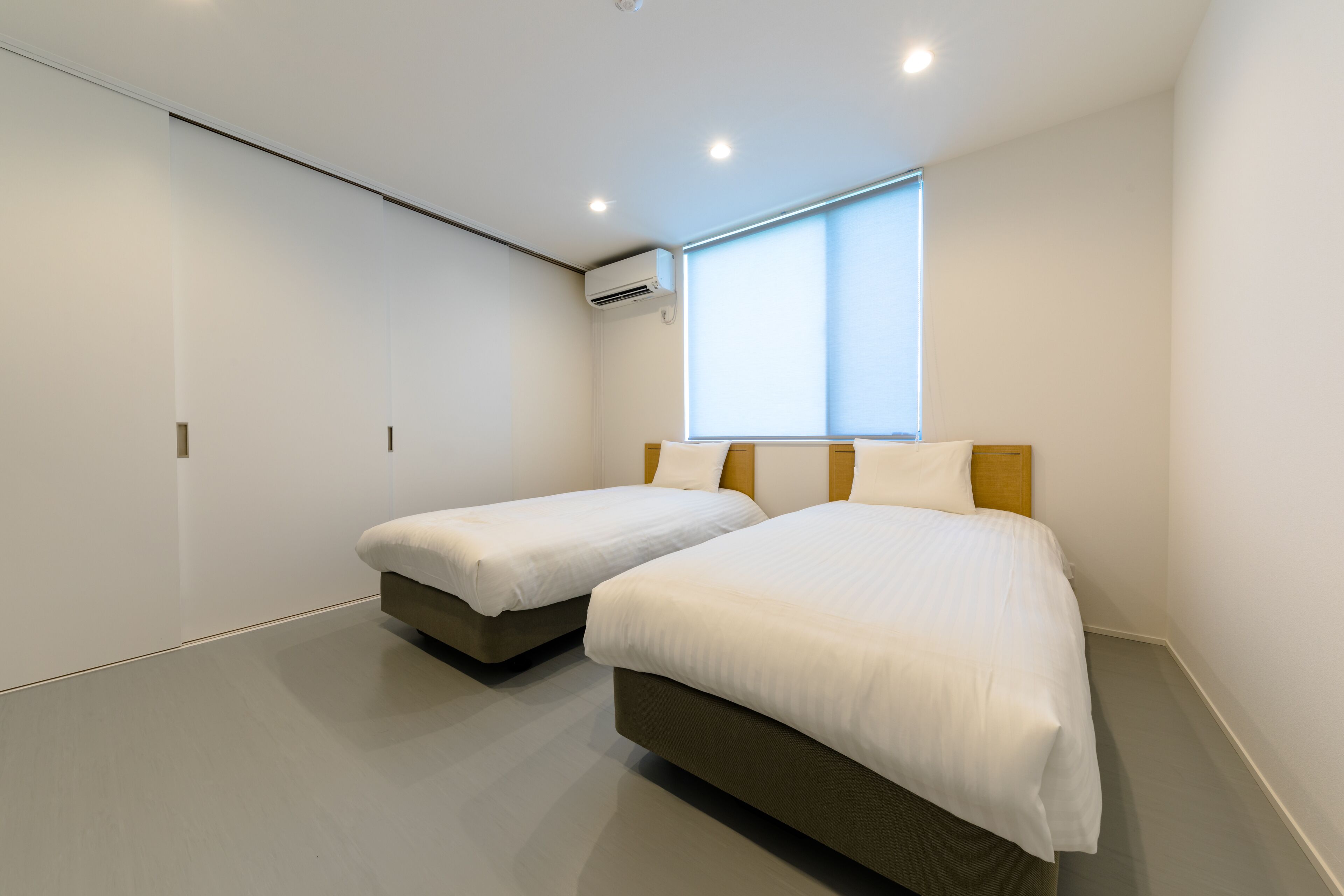 A modern hotel room with two beds, a large window, and an air conditioning unit.
