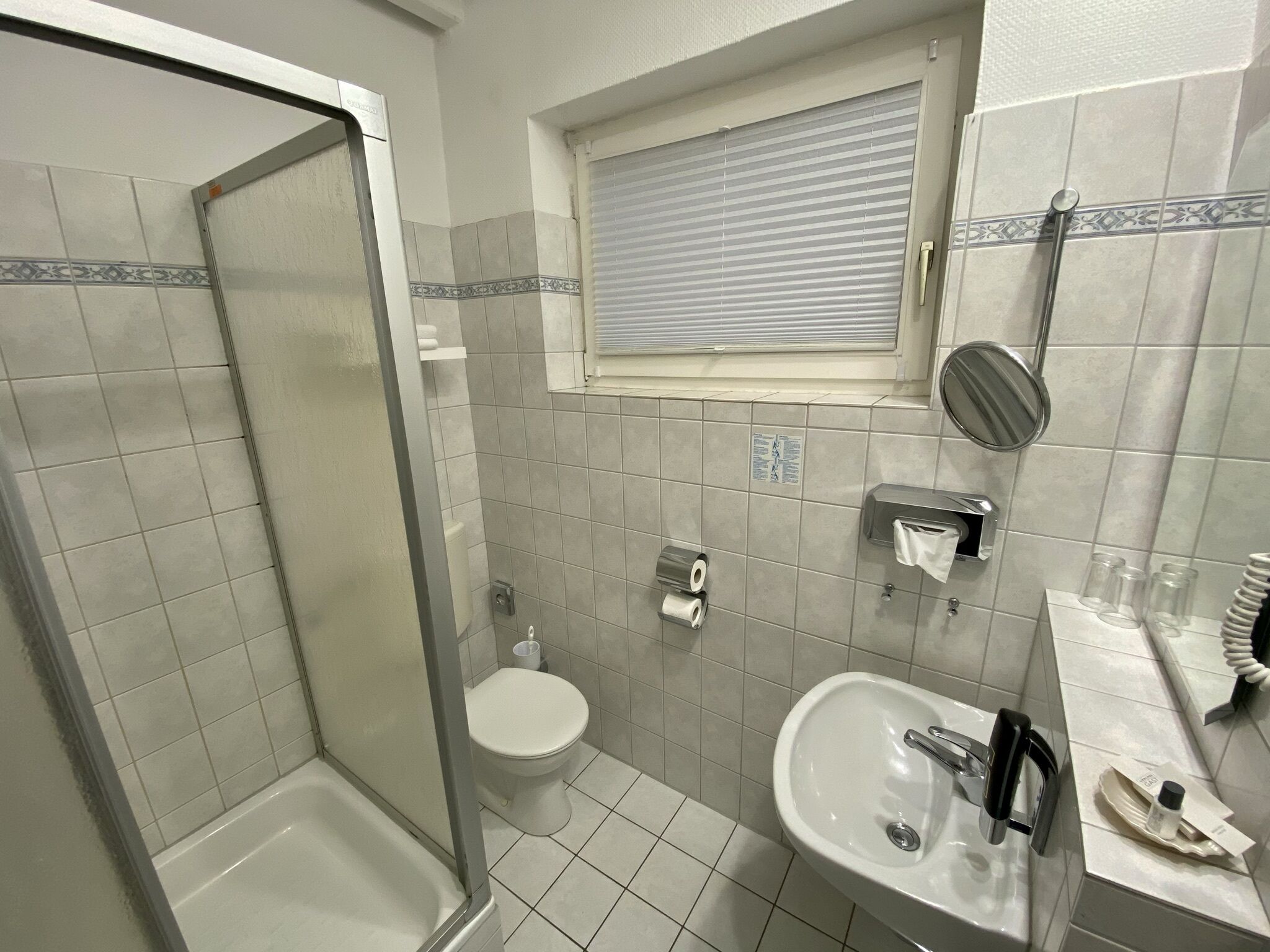 A bathroom with a shower, toilet, sink, and a window with blinds.