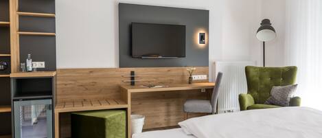 A modern hotel room with a bed, a desk with a flat-screen TV, a green armchair, and a built-in mini-fridge.