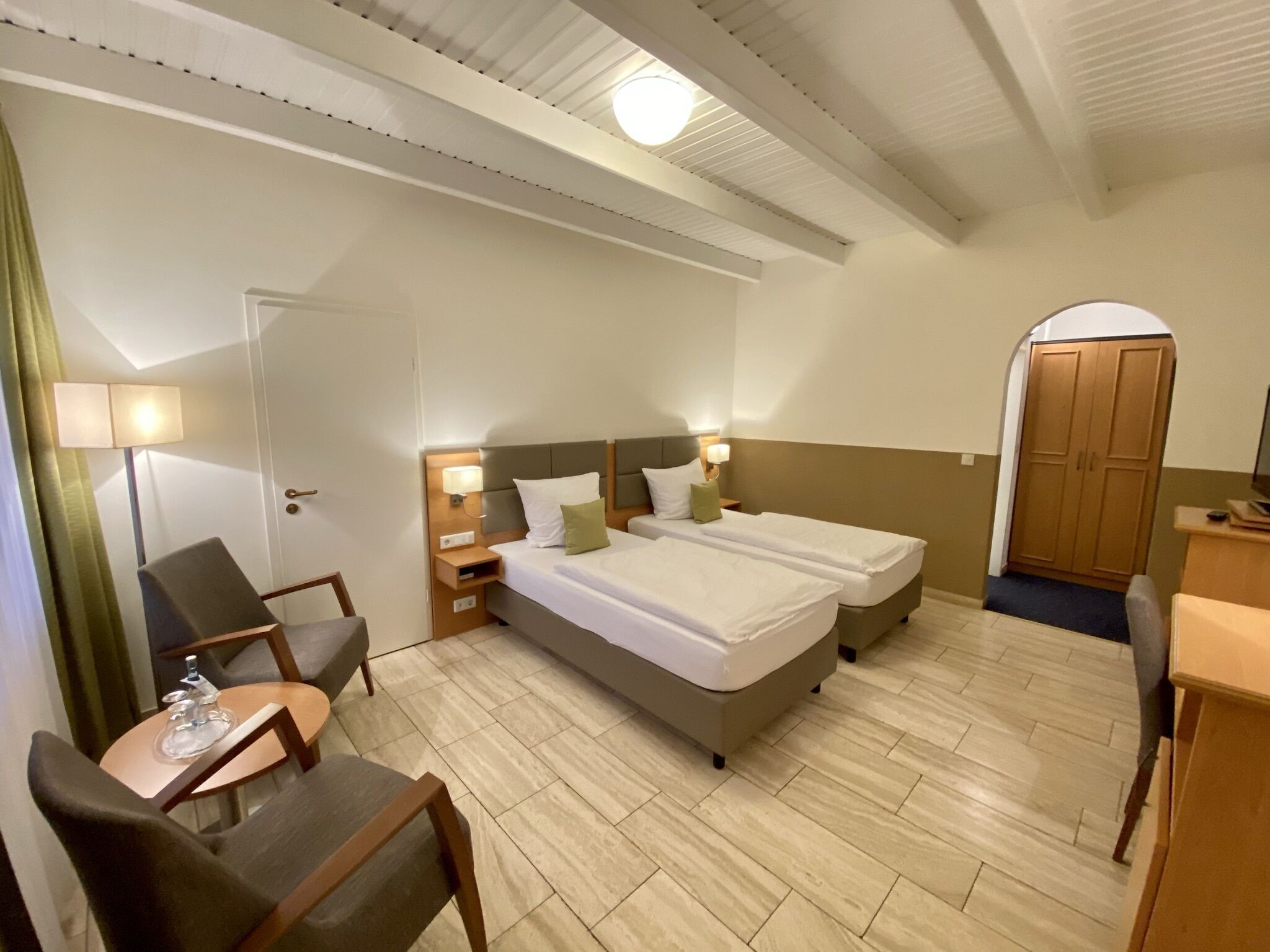 A hotel room with a double bed, two bedside tables with lamps, a desk with a chair, a wardrobe, and a door leading to another room.
