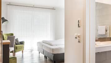 A modern hotel room with a bed, a green armchair, a desk, and a bathroom with a sink and mirror.