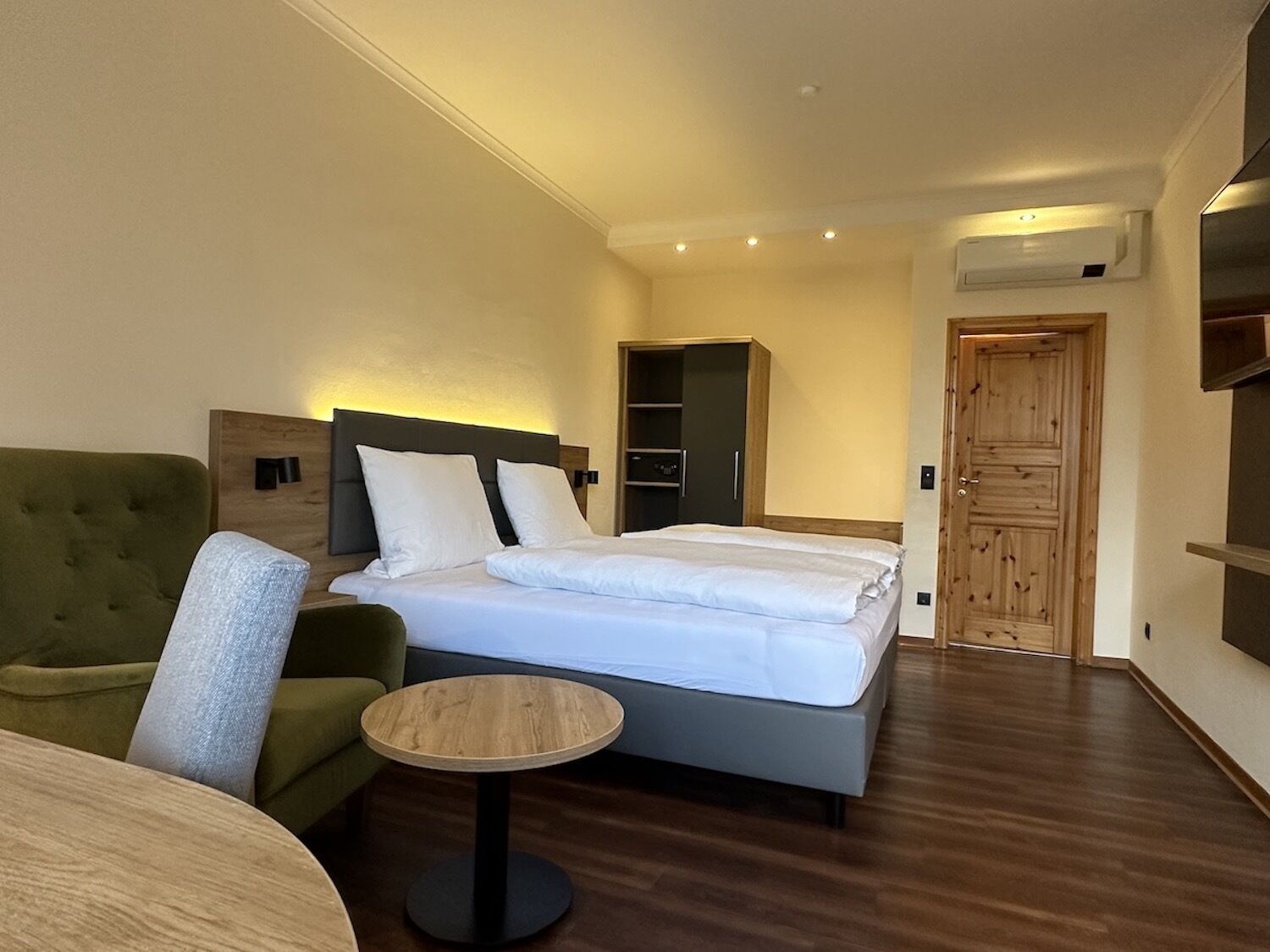 A hotel room with a bed, a green armchair, a small round table, and a wooden wardrobe.
