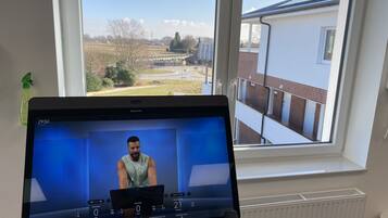 Een Peloton hometrainer met een scherm waarop een trainingssessie wordt weergegeven, staat voor een raam met uitzicht op een woonwijk.