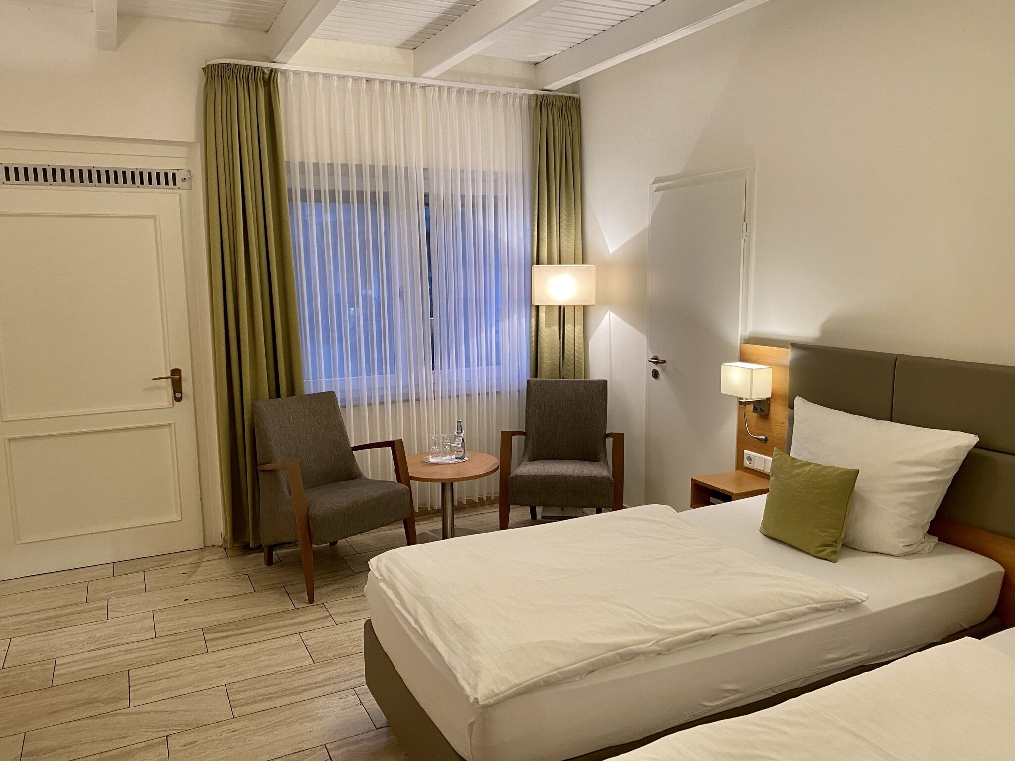 Hotellihuone, jossa on sänky, kaksi nojatuolia, pieni pöytä, lamppu ja ikkuna, jossa on ohuet verhot.
