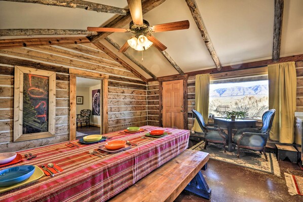 Interior d'una cabana rústica amb una taula de dinar preparada per a quatre, parets de fusta, un ventilador al sostre i vista a les muntanyes per una finestra.