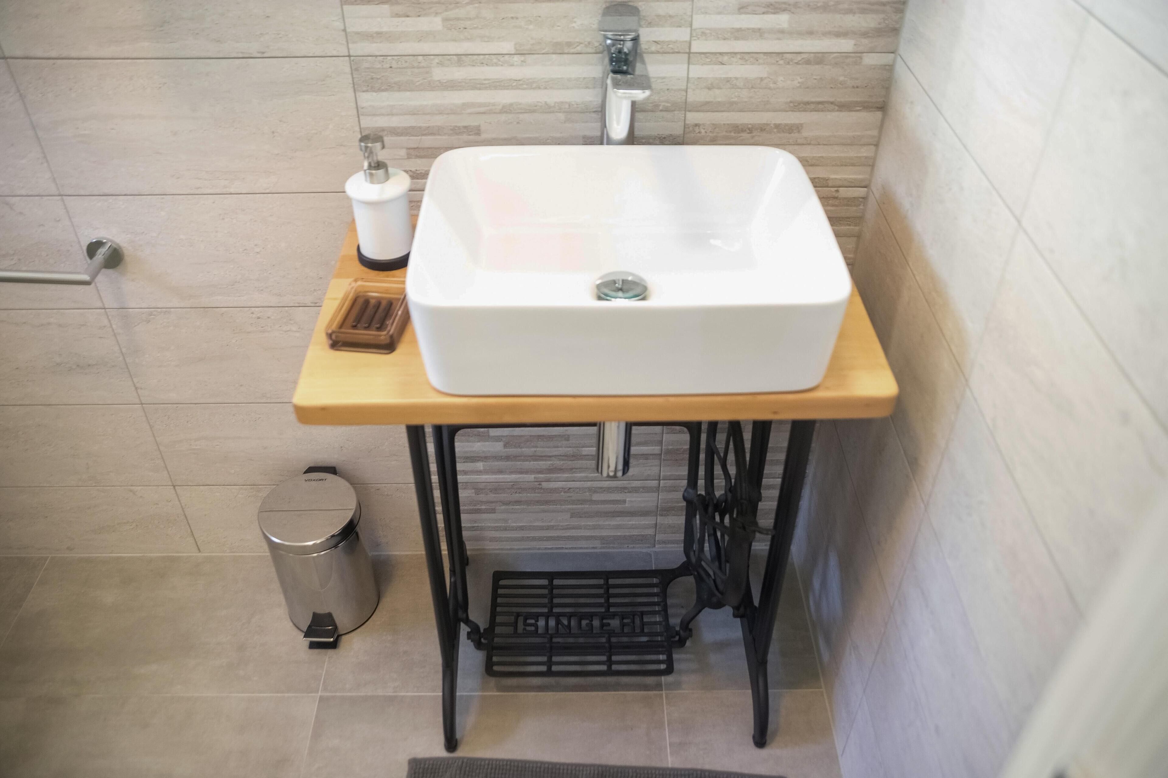 A bathroom with a white sink on a wooden vanity, a stainless steel trash can, and a tiled floor.