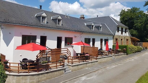 Ein Gebäude mit heller Fassade, mehreren Fenstern und einer überdachten Außenbereich-Sitzgelegenheit mit roten Sonnenschirmen.