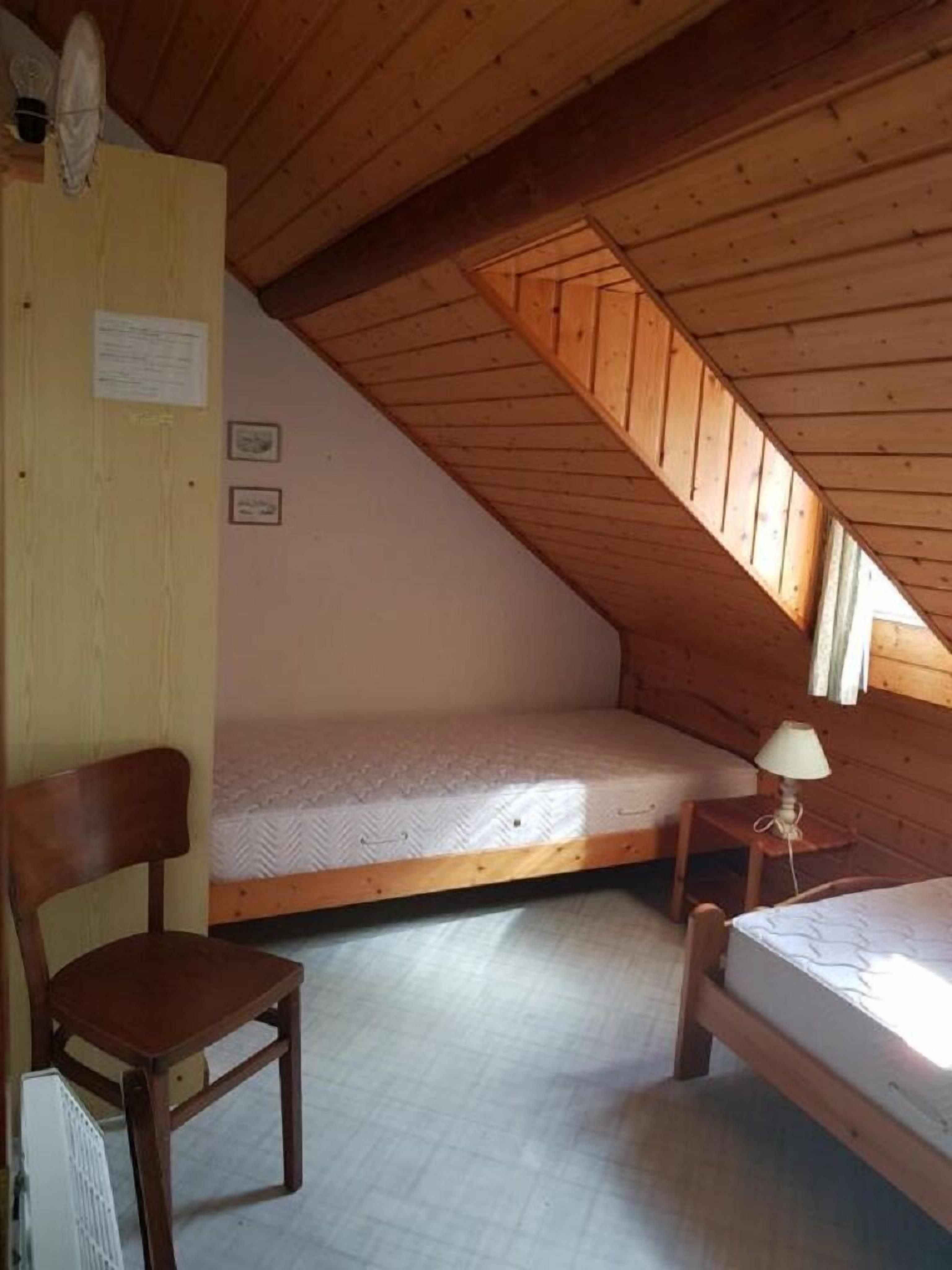 A small, single-room cabin with a sloped ceiling, a wooden chair, two single beds, and a small table with a lamp.