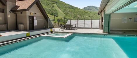 Una piscina con vista a una montaña, un área techada con mesas y un edificio con un cartel.