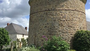 Ein steinerner Turm mit einer spitz zulaufenden Haube, umgeben von Grün und einem weißen Gebäude mit grauem Dach im Hintergrund.