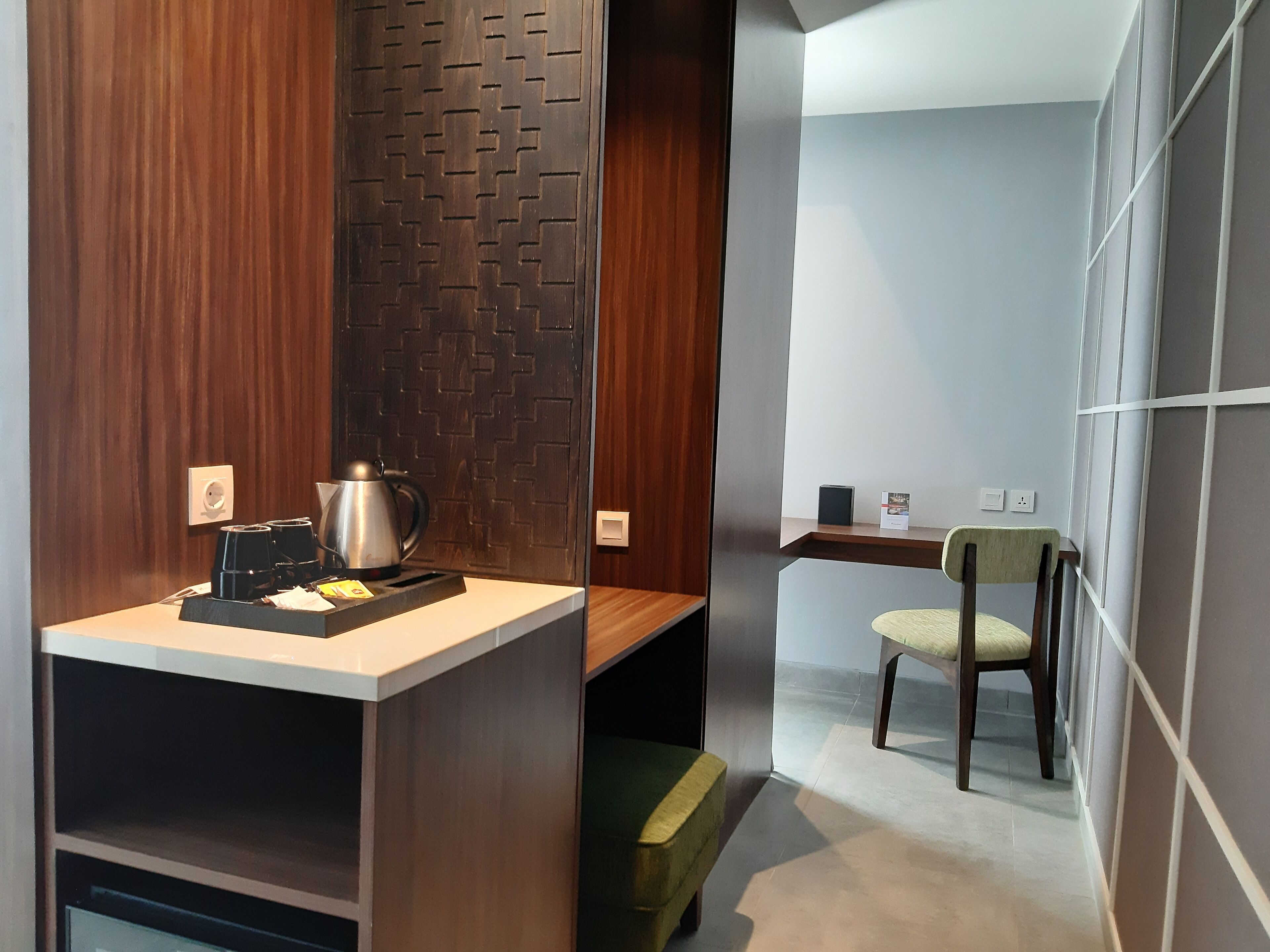 A modern hotel room with a wooden accent wall, a small desk with a kettle, a chair, and a shelf with a framed picture.