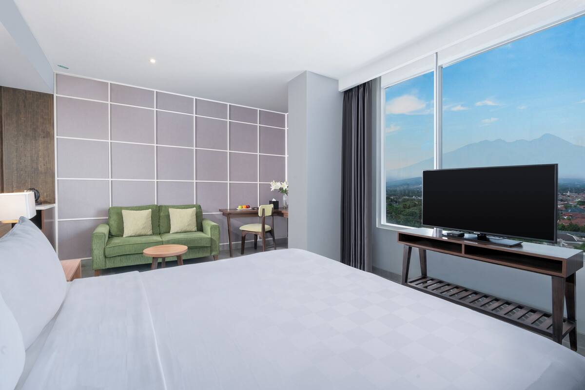 A hotel room with a large bed, a green sofa, a small table, a TV on a stand, and a city view through a large window.