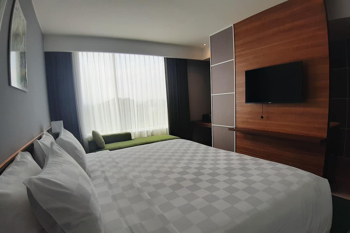 A hotel room with a large bed, a TV mounted on the wall, a window with sheer curtains, and a green upholstered bench.