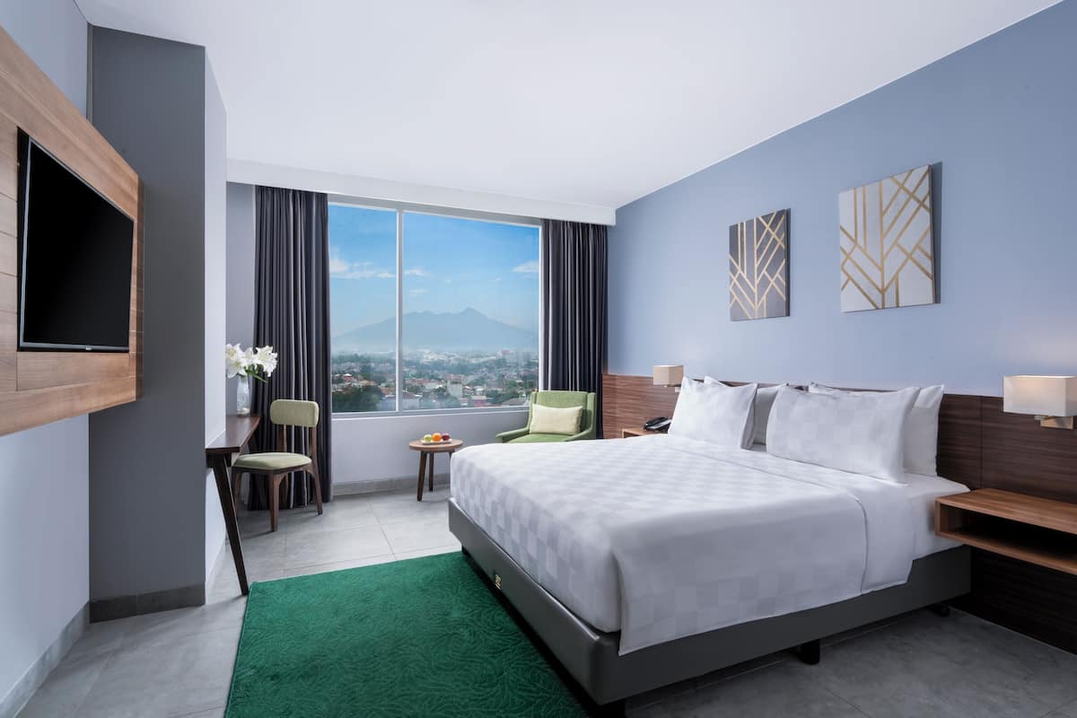 A modern hotel room with a large bed, a desk, a chair, a TV, and a view of a cityscape through a large window.