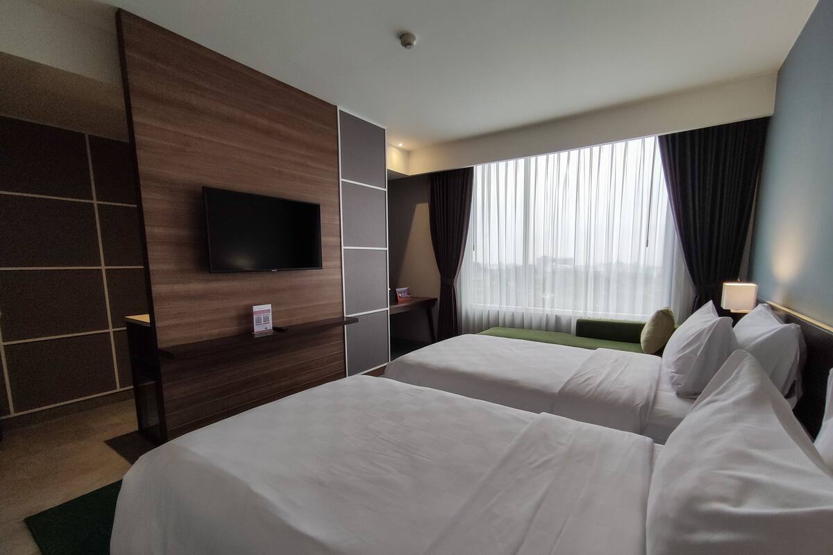 A hotel room with two beds, a flat-screen TV mounted on the wall, a window with sheer curtains, and a bedside table with a lamp.